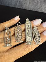 Four Egyptian sterling silver hieroglyphic pendants held on fingers showing front face design and size.