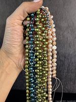 Hand holding seven strands of genuine coloured pearls in green, blue, cream and light pink. Loose string ends visible.