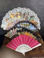 Six vintage hand fans arranged overlapping, showing intricate lace trims and painted fabric designs with bright pink fan at front