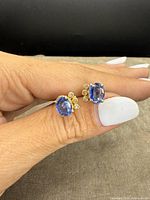 Close-up photo showing both earrings held between fingers, showing oval blue stones and three small diamonds on each earring.