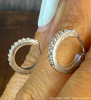 Close-up of the front and side of both sterling silver half-hoop earrings with clear stones held in a hand, showing the textured silver and embedded stones.