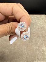 Stud earrings held in hand showing front view with blue central stones and clear stones with rose-gold overlay