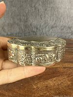 Front side view of small oval silver snuff box showing detailed floral and leaf decoration on raised metalwork around the edge.