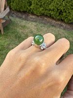 Ring worn on finger showing smooth polished green jade sphere and silver band with cluster accents, outdoor lighting.
