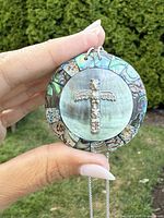 Hand holding large circular pendant showing front with sterling silver totem pole and colorful abalone inlay border against outdoor greenery background.