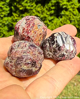 Three natural raw garnet boulder specimens held in a hand outdoors. The specimens show deep reddish tones with rough texture, various sizes visible.