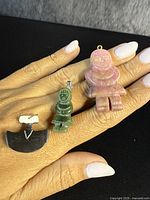 Three vintage Inuit carved stone pendants held on hand for scale, showing a pink human figure pendant with hanging loop, a green human figure pendant with silver bail, and a black whale or animal shaped pendant with silver bail.