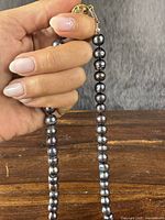 Hand holding a segment of the black pearl necklace showing the golden-toned sterling silver clasp and varying sizes of pearls.
