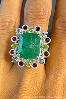 Close-up of ring on finger displaying large square emerald surrounded by red, green and blue gemstones in sterling silver setting.