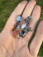 Seven faceted natural mystic topaz gemstones and one amber colored faceted gemstone displayed on a hand outdoors on grass background, showing iridescent colors.