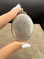 Front view of large oval Birks sterling silver locket pendant held by hand showing floral engraving design