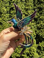 Brooch held in hand showing front side with colorful enamel in green, blue, and purple and clear rhinestones on body and tail.