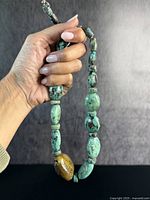 Hand holding a turquoise and brown boulder beaded necklace against a dark background to show size and detail.