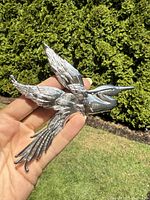Hand holding a large silver-tone bird brooch outdoors showing detailed feathers and three-dimensional shape.