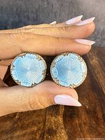 Front view of both clip-on earrings held between fingers, showing pale blue guilloche enamel and detailed sterling silver edges.