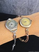 Photo showing both antique ladies wrist watches worn on wrist, one with silver engraved case and black braided strap, the other with smooth silver case and brown braided strap.