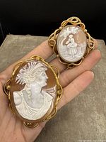 Photo of both antique cameo brooches held in hand showing front carved shell details and gold-tone frames
