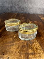 Two early antique French brass mounted glass dresser boxes side by side on a wooden table under soft lighting, showing top and sides with oval shapes and brass rims.