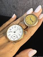 Two antique trench-style wrist watches shown on finger for scale, one with a white dial and the other yellowed dial, both showing case and crown details.