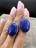 Close-up of one large sterling silver earring with natural lapis lazuli oval stone held between fingers showing front face.