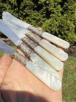 Photo showing five butter spreaders held in hand, highlighting mother of pearl handles and sterling silver mounts with engraved detailing.
