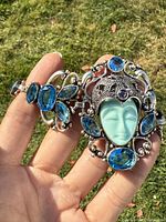 Bracelet held in hand outdoors showing detailed front with turquoise carved face and blue gemstone accents.