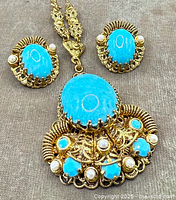 Close-up detail of necklace pendant and matching earrings showing large bright blue glass stones, smaller blue glass stones, and faux pearls set in gold-tone filigree frames.