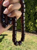 Hand holding cherry red beaded prayer necklace showing bead shape and color with outdoor greenery background.