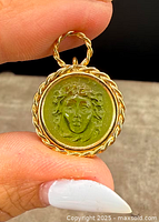 Close-up front view of the Medusa pendant showcasing the detailed face on a green base with gold overlay frame and bail held between fingers.