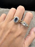 Close-up of two gold-tone rings on fingers, showing the dark blue gemstone surrounded by clear stones and the clear gemstone with smaller stones.