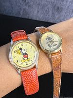 Two vintage Mickey Mouse wristwatches shown on wrist; one with red-orange textured strap, gold-tone case and colored Mickey on dial; second with light brown leather strap and gold-tone dial with Mickey figure.