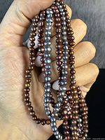 Close-up of multiple strands of genuine pearls in bronze, copper, and blue-grey tones held in hand, showing color and texture.