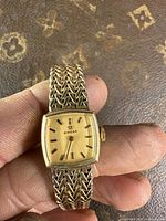 Front view on hand showing gold-tone square dial and woven mesh bracelet.