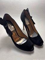 Full side angle view of the pair of black satin open toe high heels with bows at the back.