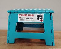 Closed collapsible stool showing its turquoise color, white top grips, and brand label indicating it holds up to 100 kg.