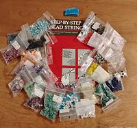 Top-down view of assorted beads arranged on a table around a red instructional book titled 'Step-By-Step Bead Stringing.' Multiple small resealable bags holding various colors and styles of beads are visible.