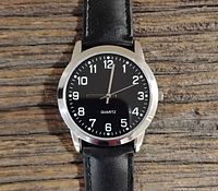 Close-up of men's quartz watch with black dial, white numerals, silver hands, and black leather strap on wooden surface.