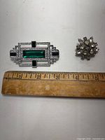 Two rhinestone brooches side by side with a ruler for scale, shows rectangular green rhinestone brooch and smaller clear rhinestone cluster brooch.