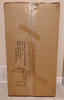 Photo of unopened cardboard box containing space saving desk in flat pack form. Box shows printed diagram of desk and item specifications.