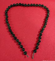 Full view of the entire necklace displayed on a red textured surface showing 47 dark green polished beads strung on green thread.