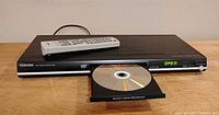 Front view of Toshiba DVD player with disc tray open and remote on top.