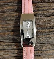 Close-up of the watch face showing Nina branding, numeric markers 12 and 6, rectangular silver-tone case and pink leather strap.