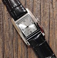 Close-up photo of women's quartz watch with rectangular silver-tone case and black leather strap on wood background.