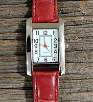 Close-up image showing the watch face with rectangular silver-tone casing, white dial, black numbers, and red second hand.
