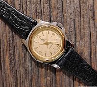 Close-up angled view of the women's SK quartz watch showing the gold-tone dial, silver-tone case with gold bezel, and black textured leather strap on wooden surface.