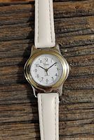 Close-up photo of the round silver-tone Timex quartz watch face with white dial, black Arabic numerals, black hour and minute hands, and a silver second hand on a wooden background.