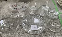 Top view of 10 clear glass and crystal items arranged on carpet: pedestal bowl with three spheres inside, two shallow bowls, two small candle holders, fluted bowl, divided plate, and multiple textured serving plates.