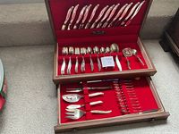 Photo showing full 80-piece stainless steel silverware set in a wooden case with red lining, organized in two layers: knives on the top layer lid, various forks and spoons below them, and additional serving utensils in the bottom drawer.