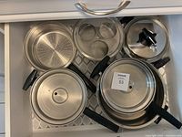 Overview of the aluminum pots and pans set in a drawer, showing various pots, pans, lids, and steamer inserts with black handles.