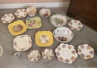 Overview of assorted decorative plates and bowls with various floral and fruit patterns in multiple colors including white, yellow, and multicolored designs shown together on floor.